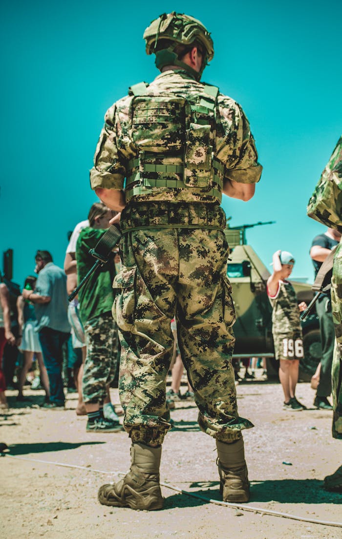 A soldier in military camouflage standing outdoors, surrounded by people and vehicles.