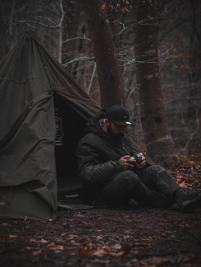 pexels photo 14115122 A man sits by his tent in a dark, moody autumn forest, capturing the essence of camping adventure.