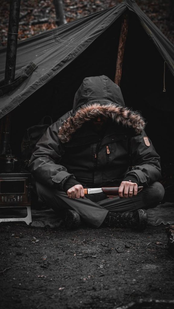 A camper in a hooded jacket holding a knife outside a tent in a dark forest setting.