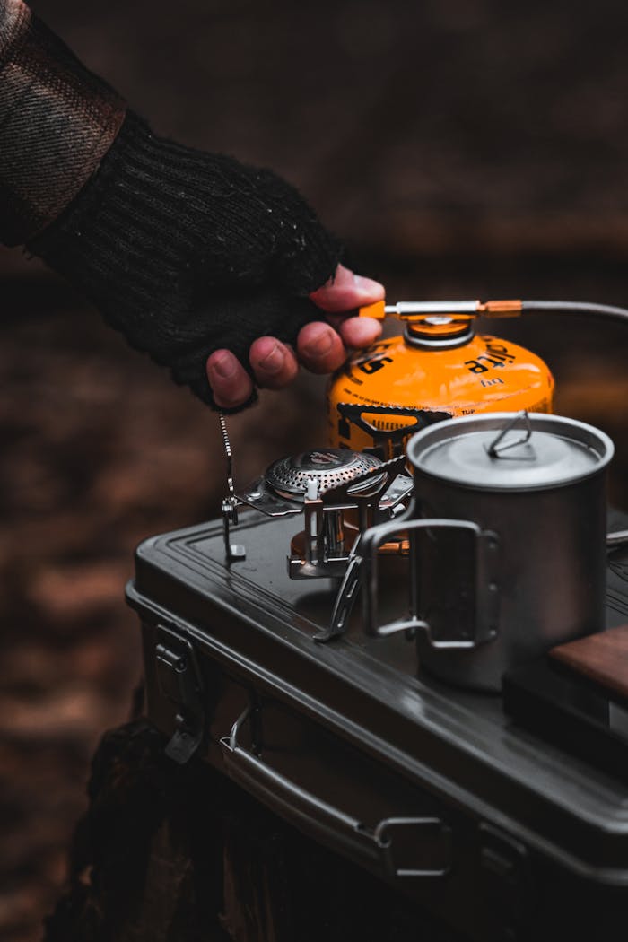 A hand in a glove uses a portable camping stove outdoors, illustrating adventure and travel.