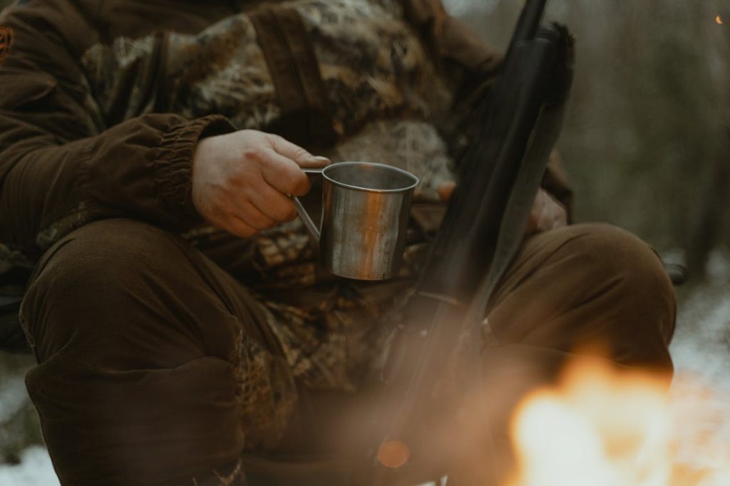 pexels photo 6204613 A person in camouflage enjoys a campfire with a tin cup outdoors.
