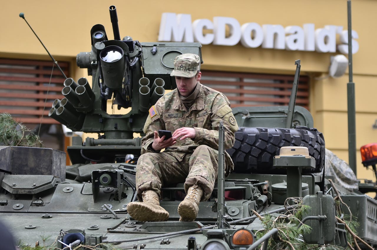 A soldier in camouflage resting on an armored vehicle, using a smartphone outside a McDonald's.